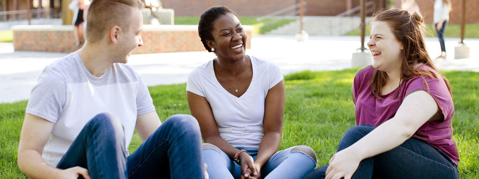 Two female students 和 one male student sitting on grass on forum laughing